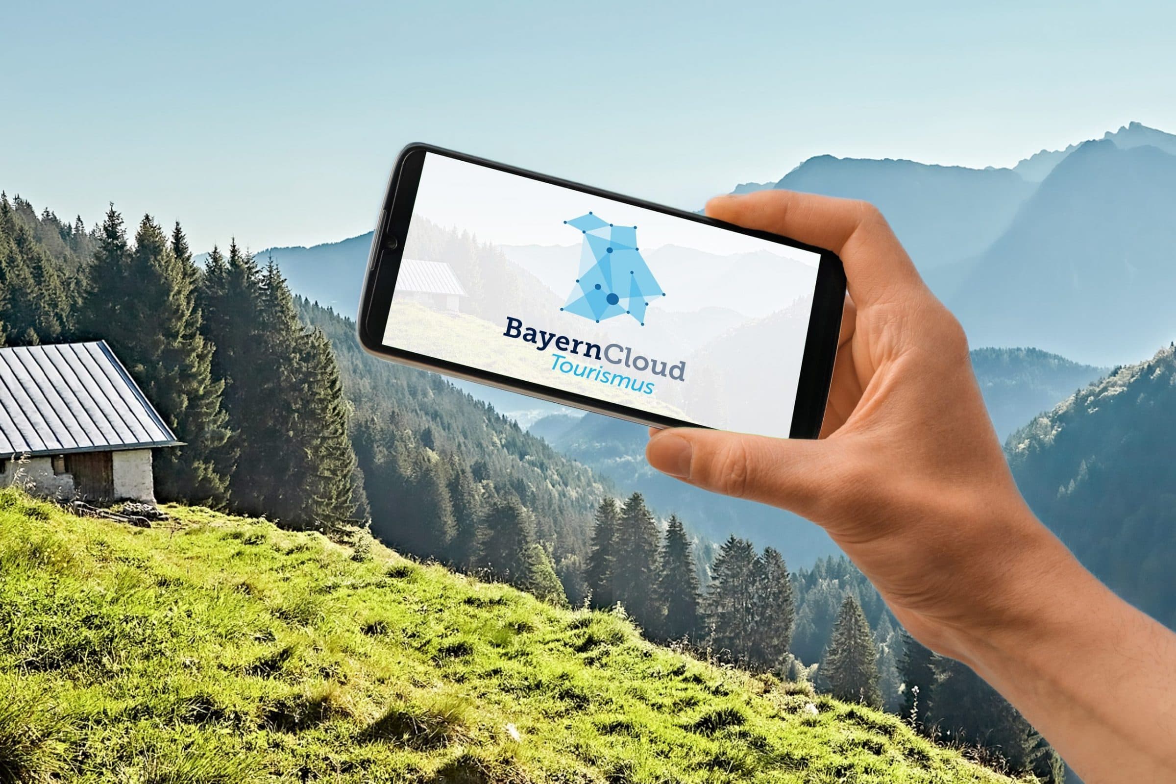 Smartphone mit dem Logo der BayernCloud Tourismus vor einer Almlandschaft